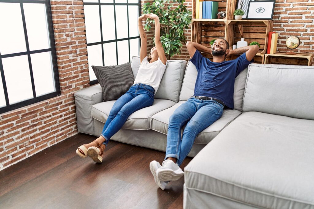 A woman and man relaxing on a couch with their arms behind their heads.